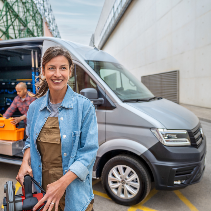 Eine Handwerkerin steht vor einem VW Crafter, in der Hand trägt sie eine Kreissäge. Im Laderaum des VW Crafter verstaut ein Handwerker Material in Kisten.