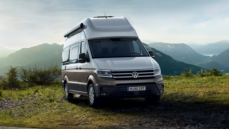 Un van Volkswagen Grand California bicolore (gris clair et marron foncé) est garé dans un paysage montagneux verdoyant, avec des collines et un ciel clair en arrière-plan.