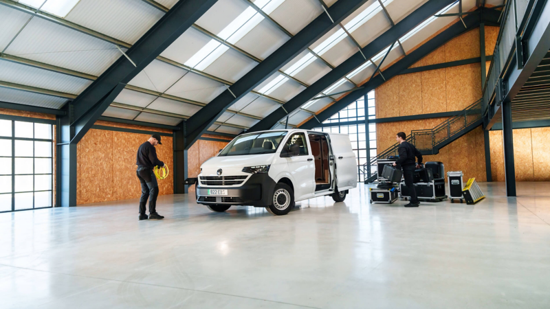 A white van in a spacious warehouse with two workers. One carries a large cable, the other handles equipment cases. Bright, industrial setting.