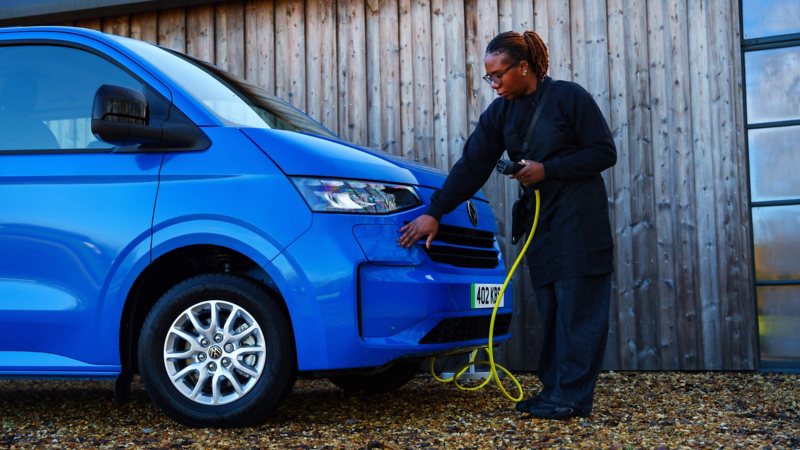 A person in dark clothing inspects a blue electric VW e-Transporter van, holding a yellow charging cable. The scene conveys focus and innovation in eco-friendly transport.