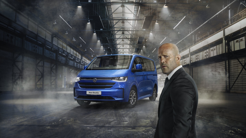 Jason Statham standing in front of the new VW Transporter Shuttle parked in a warehouse setting. 