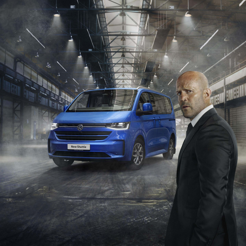 Jason Statham standing in front of the new VW Transporter Shuttle parked in a warehouse setting. 