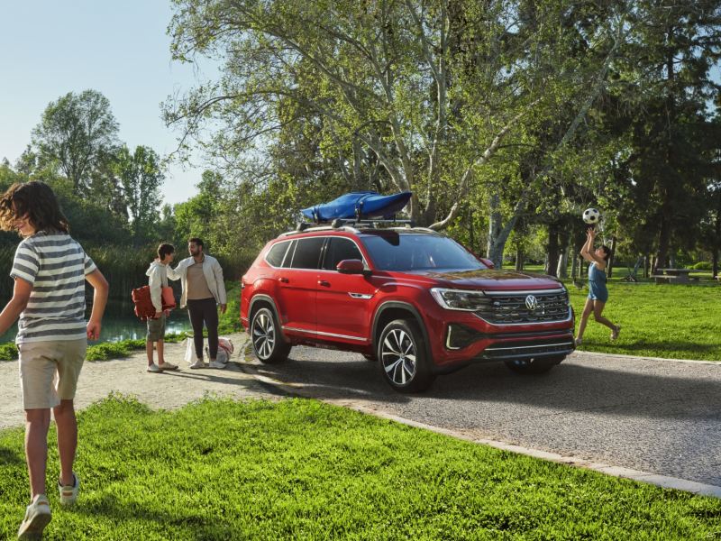 The 2024 Aurora Red Chroma Volkswagen Atlas with a canoe attached at the roof parked on the road in the park.