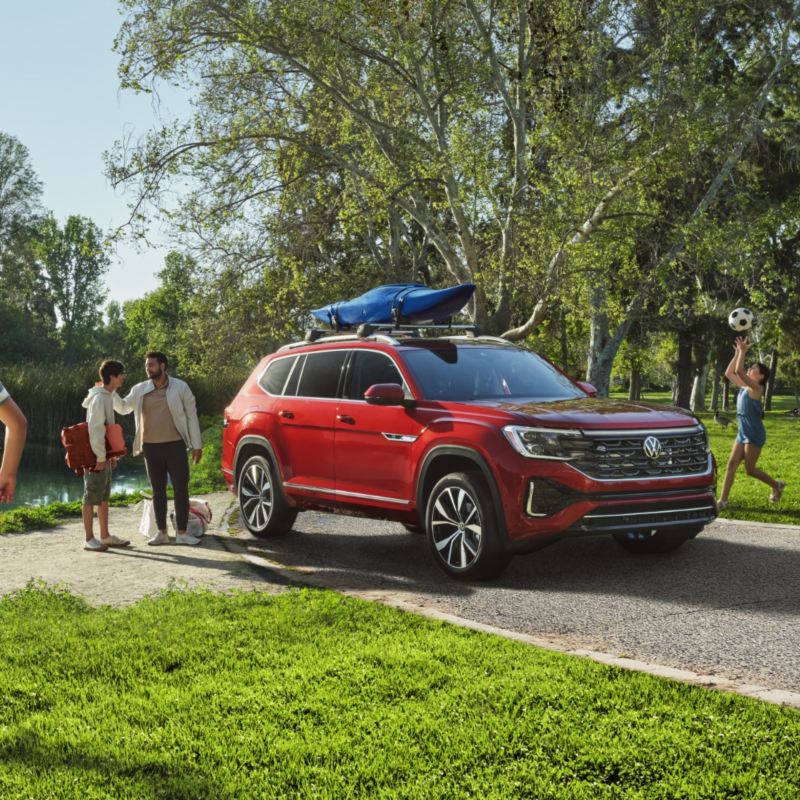 The 2024 Aurora Red Chroma Volkswagen Atlas with a canoe attached at the roof parked on the road in the park.