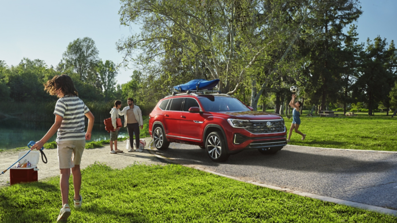 The 2024 Aurora Red Chroma Volkswagen Atlas with a canoe attached at the roof parked on the road in the park.