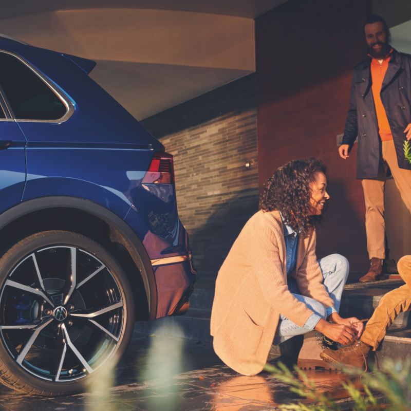 A woman helping a kid to tie his shoe next to the Blue Metallic Volkswagen SUV