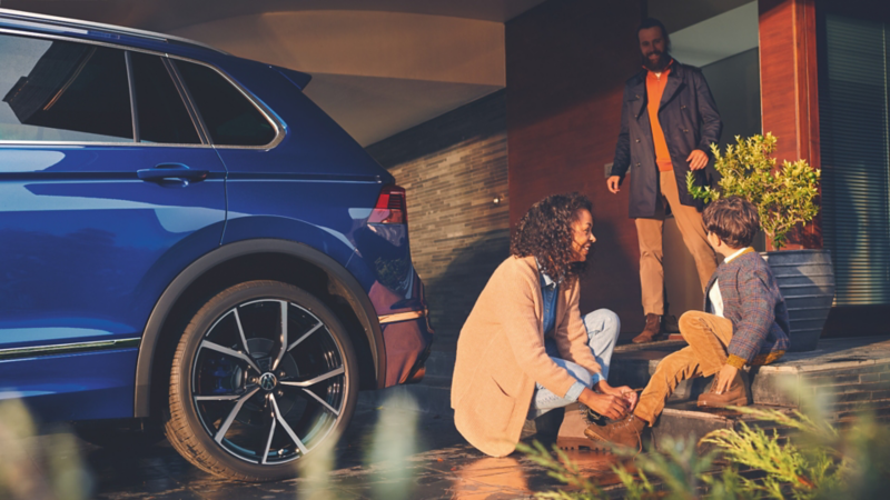 A woman helping a kid to tie his shoe next to the Blue Metallic Volkswagen SUV