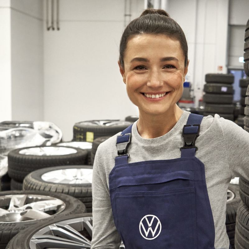 Une technicienne Volkswagen qui sourit dans un garage, avec beaucoup de pneus en arrière-plan.