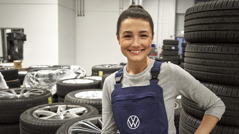 Une technicienne Volkswagen qui sourit dans un garage, avec beaucoup de pneus en arrière-plan.