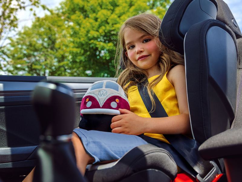 Une petite fille assise dans un siège pour enfant dans une voiture qui tient un jouet de véhicule Volkswagen dans sa main.