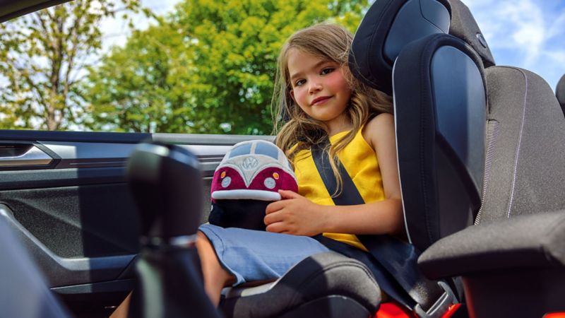 A small girl sitting in the kid’s chair in the car and holding a Volkswagen toy in her hand