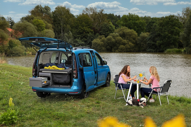 Vista posteriore di Nuovo Caddy California Volkswagen, parcheggiato vicino a un lago, con il portellone posteriore aperto e la cucina a scomparsa in utilizzo. Due ragazze pranzano lì accanto sedute al tavolino da campeggio.