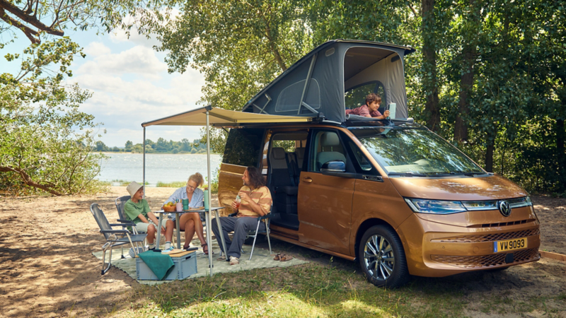 Van Volkswagen California Beach Tour stationné au bord d’un lac, toit relevable ouvert, avec des personnes installées sous un auvent à côté d’un espace pique-nique.