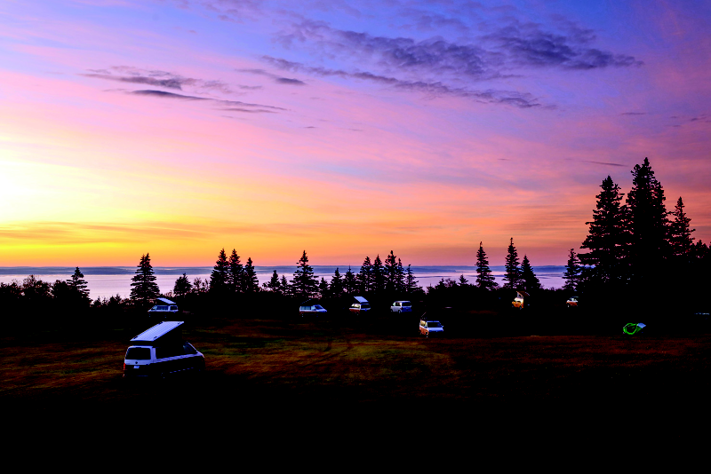 Mange biler parkeret på en eng i solnedgang med en sø i baggrunden