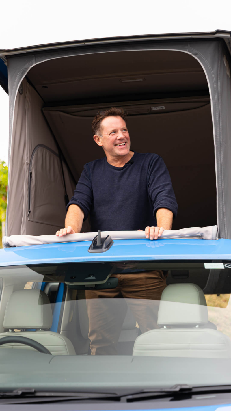 Matt Tebbutt leaning from the pop-up roof area of the camper van, smiling against an outdoor backdrop.