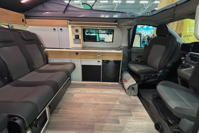 Vue de l'intérieur d'un California. Sur la gauche se trouve une banquette et un meuble de cuisine avec des finitions en bois et un réfrigérateur. Le sol est de style parquet. Le toit est relevé, et les sièges du conducteur et du passager sont pivotés vers l'arrière