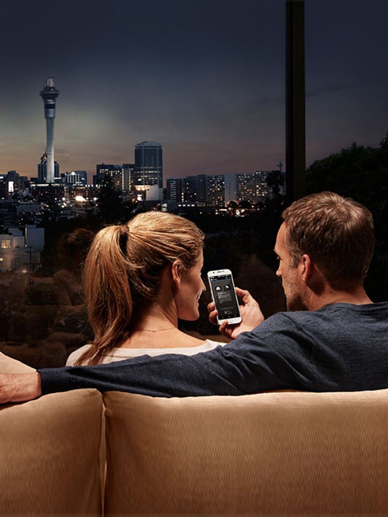 uomo e donna consultano lo smartphone seduti sul divano davanti alla finestra dove si vede città di notte