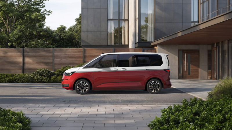 A red and white minivan is parked on a modern driveway beside a contemporary building. The scene is calm, with greenery and a wooden fence in the background.