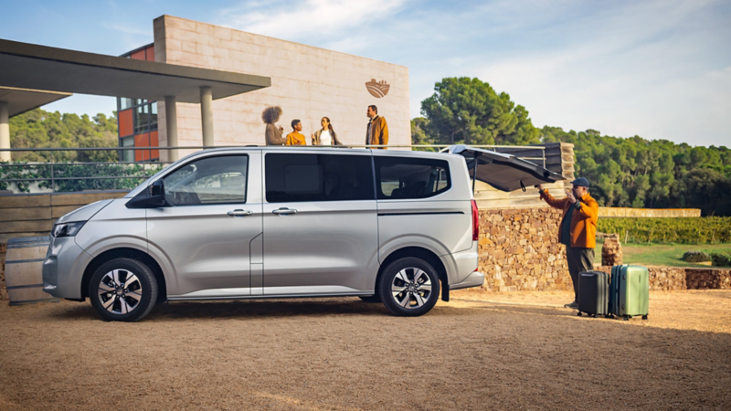 Van Volkswagen Caravelle gris stationné près d’un bâtiment, hayon ouvert, avec des bagages à côté et plusieurs personnes à proximité.