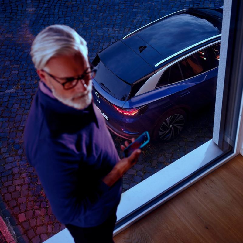 Vyras su išmaniuoju telefonu rankoje stovi prie lango, fone - mėlynos spalvos „VW ID.4“
