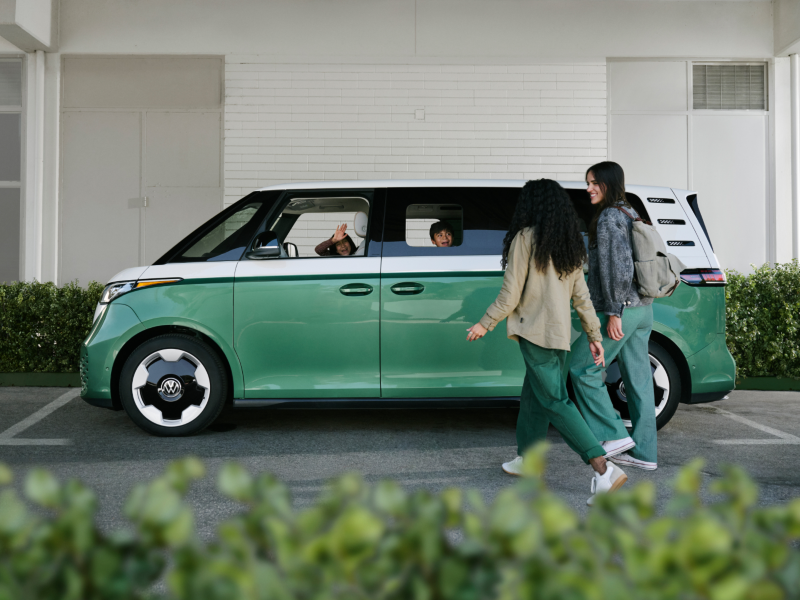 An ID. Buzz EV shown in Mahi Green Metallic and Candy White is parked in front of a building and green hedges with two children inside the vehicle happily waving to two people walking toward them.