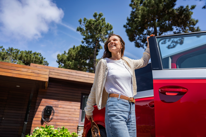 Una mujer joven sonriente y feliz, vestida con ropa casual, de pie junto a la puerta abierta de un coche rojo. Al fondo se ve una casa de madera y árboles bajo un cielo azul brillante. La escena transmite alegría y vida cotidiana.
