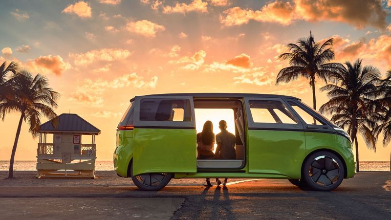 A man and woman sit inside the Volkswagen I.D. BUZZ looking out at the sunset over the ocean as the light shines through the doors and windows of the I.D. BUZZ from a side view.