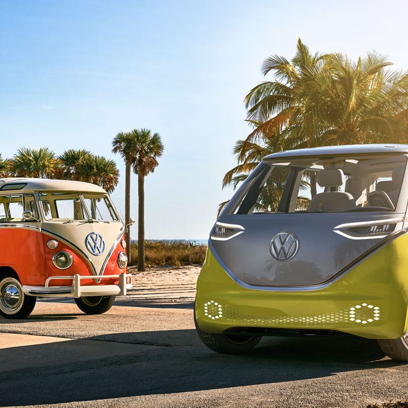Volkswagen I.D. BUZZ electric microbus and Volkswagen Samba Bus parked side by side with ocean views and palm trees in the background. 