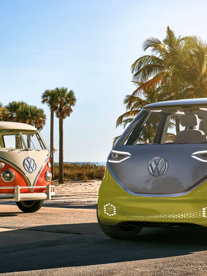 Volkswagen I.D. BUZZ electric microbus and Volkswagen Samba Bus parked side by side with ocean views and palm trees in the background. 