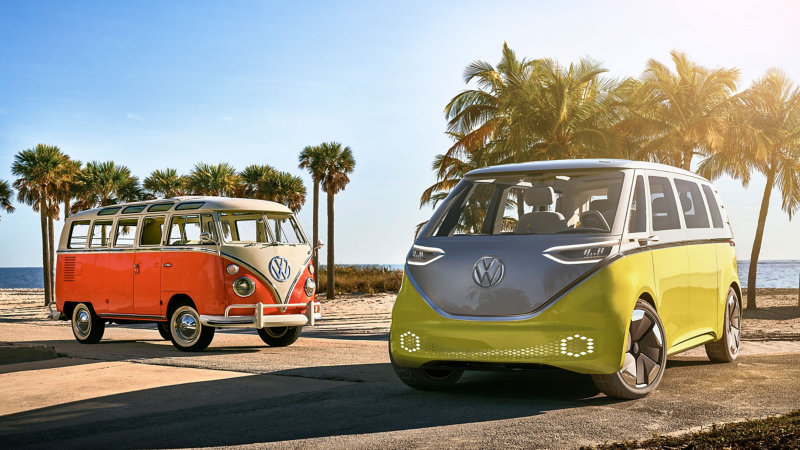 Volkswagen I.D. BUZZ electric microbus and Volkswagen Samba Bus parked side by side with ocean views and palm trees in the background. 