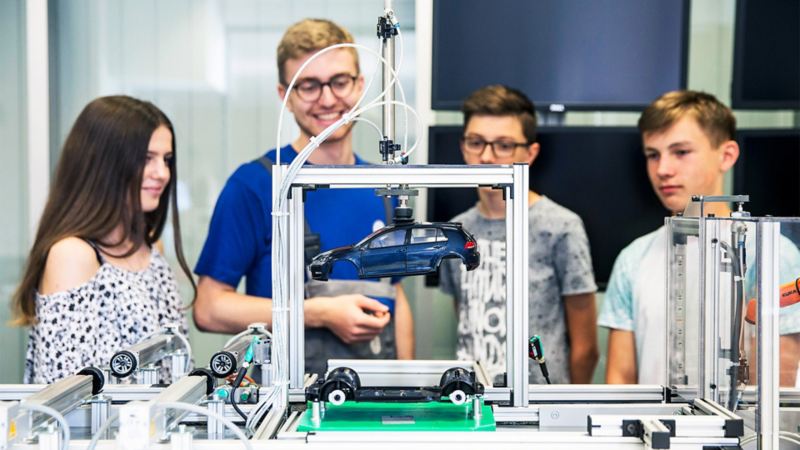 A Volkswagen employee explains vehicle production to three young people using a model