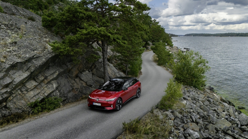 En röd Volkswagen kör längs en väg med berg på ena sidan och hav på andra