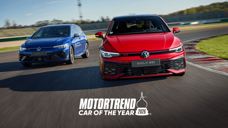 A red Volkswagen Golf GTI leads a blue Volkswagen Golf R on a race track under a clear blue sky.