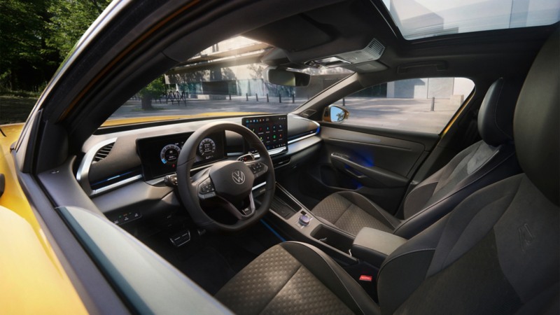  Une femme souriante est assise au volant d'une Volkswagen T-Roc. On la voit de profil, regardant vers l'extérieur du véhicule. L'intérieur de la voiture est mis en évidence, notamment les sièges en cuir noir et le toit ouvrant panoramique.