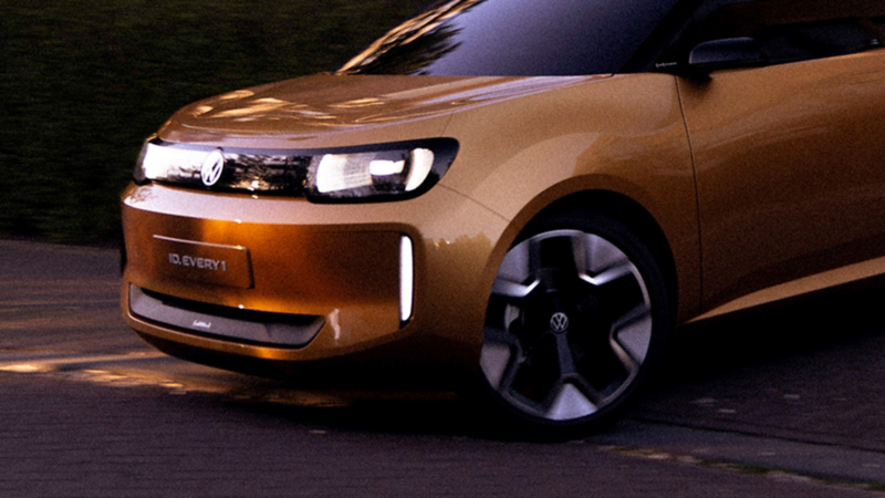 Ein goldfarbener VW ID.EVERY1 mit beleuchteten Scheinwerfen fahrend in der Dämmerung.