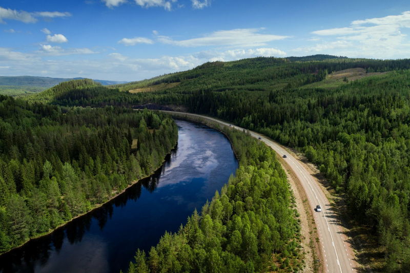 Ovanifrån ses en väg som löper längs med ett vattendrag med omkringliggande skog