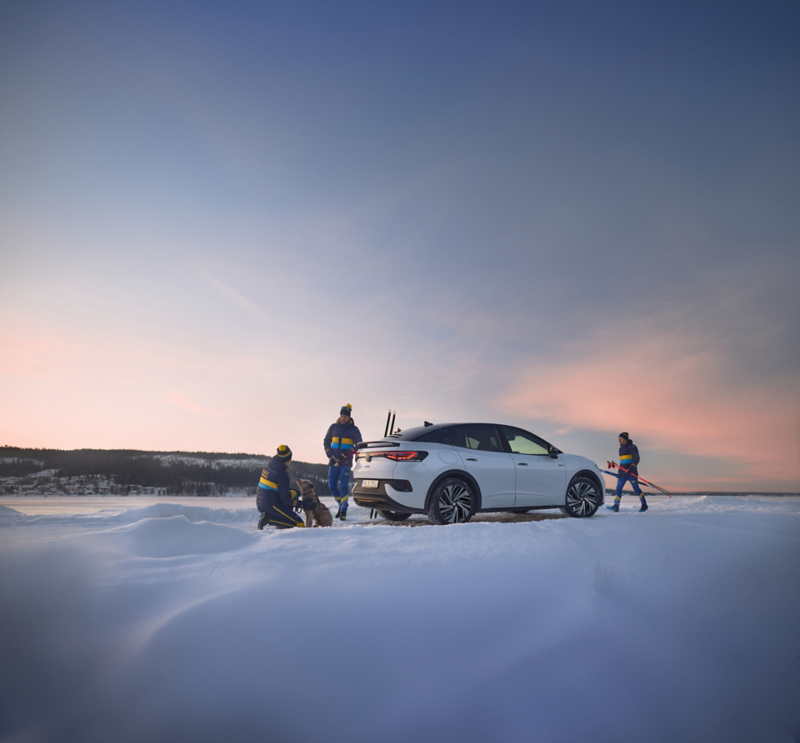 Tre personer, en hund och en Volkswagen ID.4 i vinterlandskap i en solnedgång. Fjäll och sjö i bakgrunden.