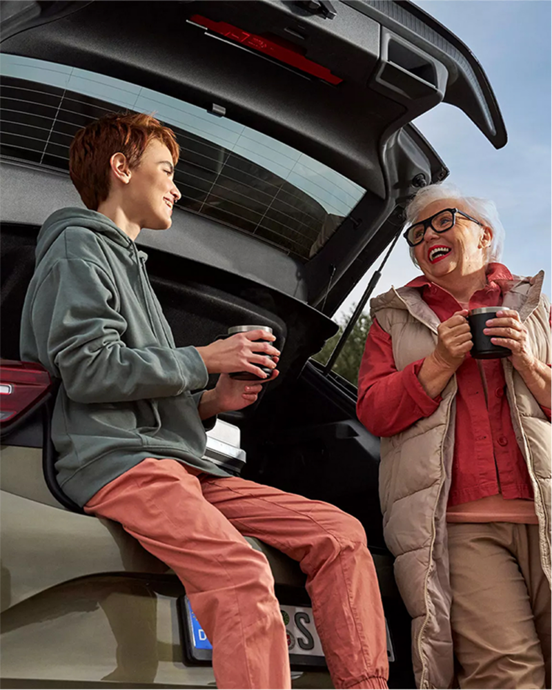 Two women enjoying a hot beverage while sitting on the tailgate of a Volkswagen vehicle.