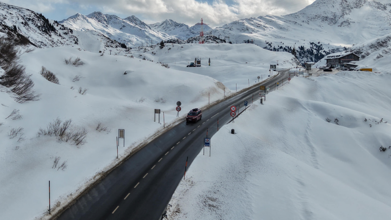 A Multivan drives on a snow-lined mountain road under a cloudy sky, surrounded by snow-covered hills and distant peaks. The scene conveys tranquility.