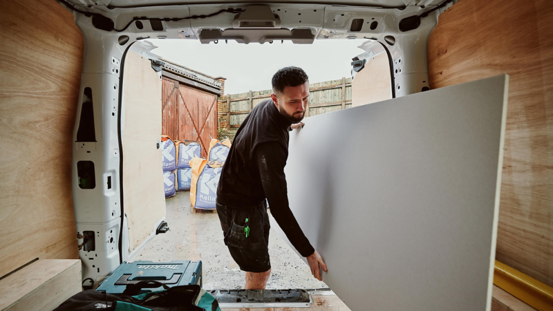 A man loads a large white panel into a van, with construction bags visible outside. The scene conveys a sense of focus and manual labor.