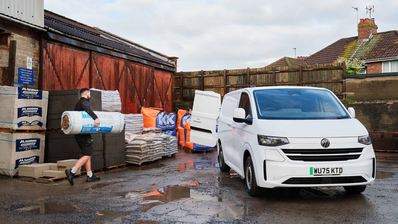 A white van with an open side door is parked beside a warehouse. A man carries a large roll of insulation. Stacks of building materials are visible. Wet ground suggests recent rain.