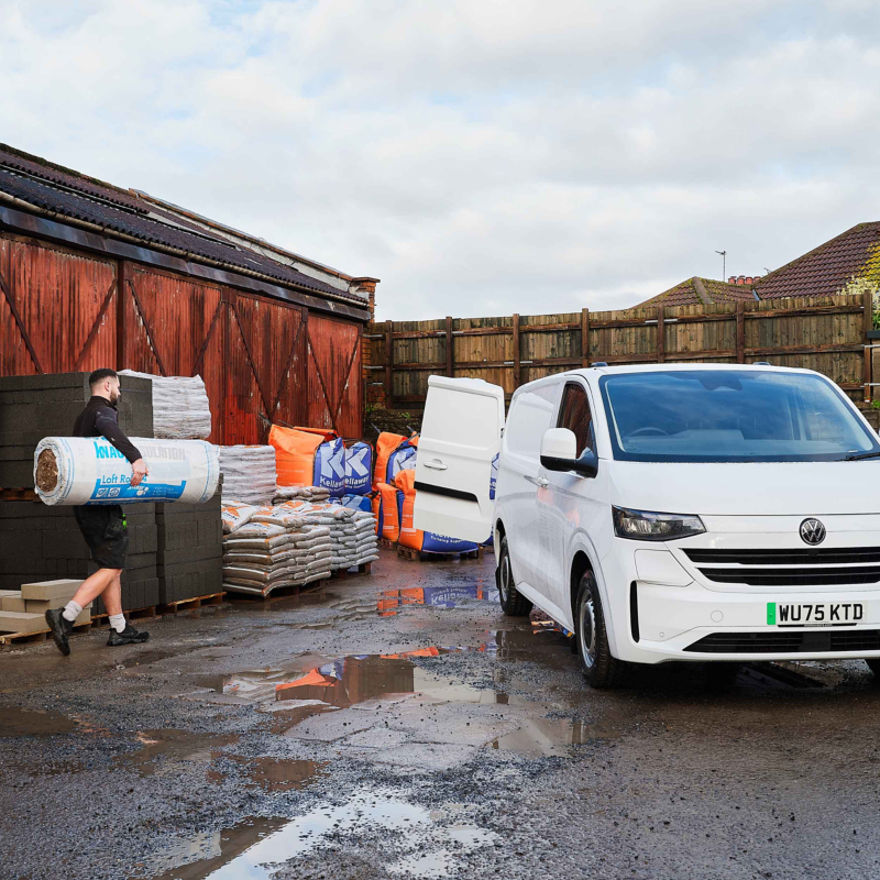 A white van with an open side door is parked beside a warehouse. A man carries a large roll of insulation. Stacks of building materials are visible. Wet ground suggests recent rain.