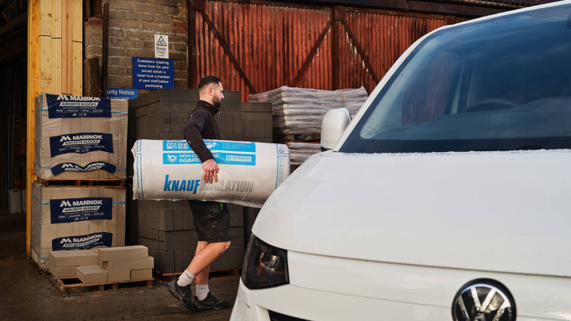 A man carries a large bag of insulation labeled "Knauf" outside a building materials store, with brick stacks and a white van in the foreground.