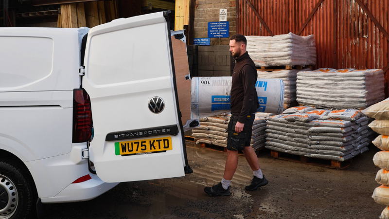 A man in work attire stands by an open white van in an industrial area, surrounded by stacked pallets of bagged goods, conveying a sense of busy activity.