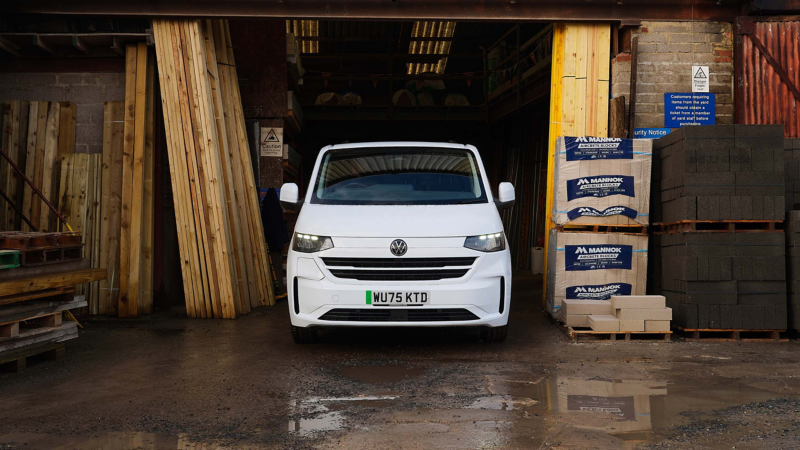 A white van is parked in a lumberyard entrance, surrounded by stacks of wooden beams and concrete bags, under a cloudy sky. The scene feels industrious.