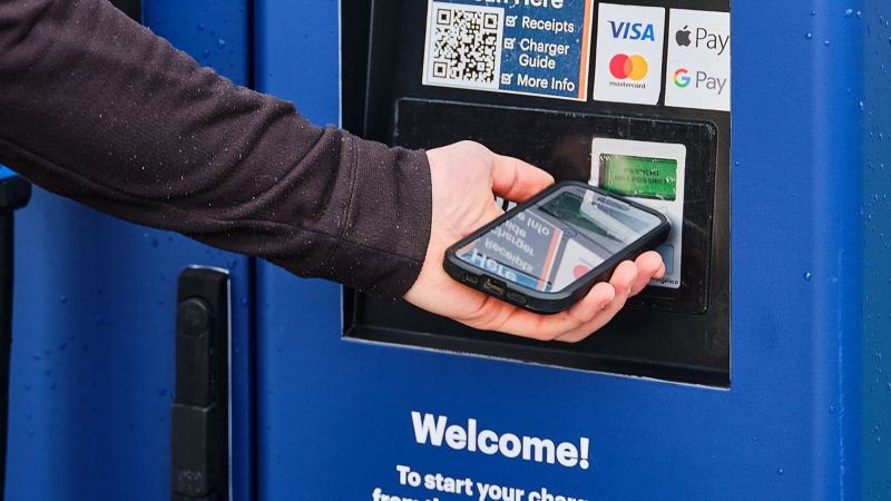 A hand holds a smartphone near a payment terminal on a blue charging station. The screen displays payment options, including Visa, Mastercard, and contactless pay.