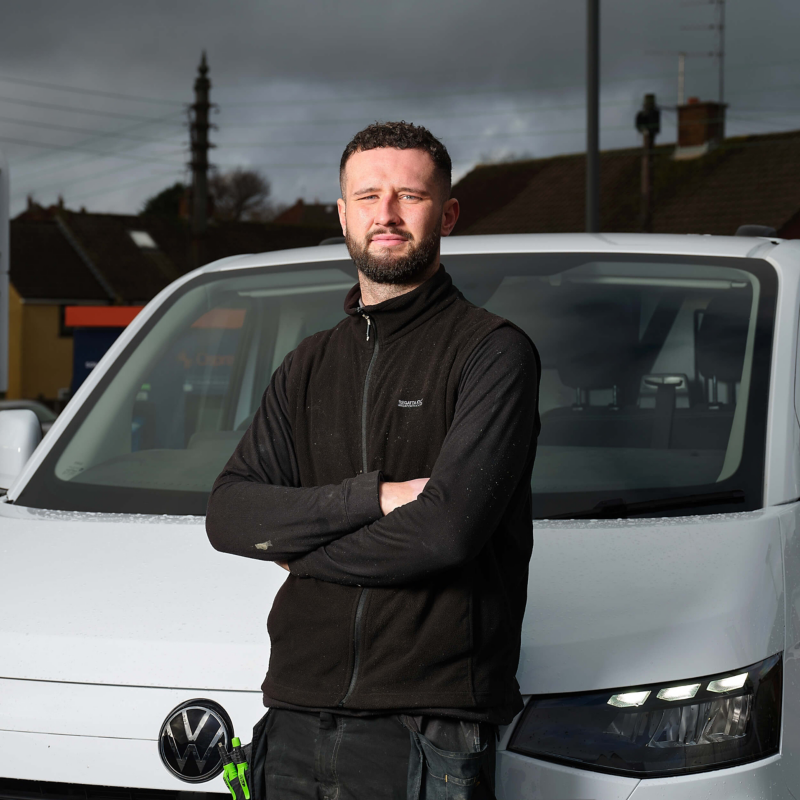 A man stands confidently with arms crossed in front of a white Volkswagen van. He wears a black jacket. Cloudy sky and rooftops in the background.
