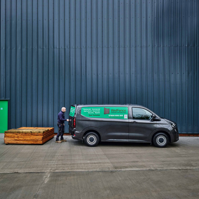 A gray van with "WellFence" and contact info is parked by a large, blue metal wall. A person is opening the van door. Stacks of wooden planks are nearby.
