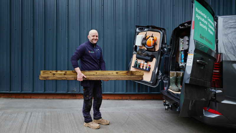 A man in work clothes holds wooden planks, standing beside an open van with neatly organized tools. He smiles against a metal building backdrop.