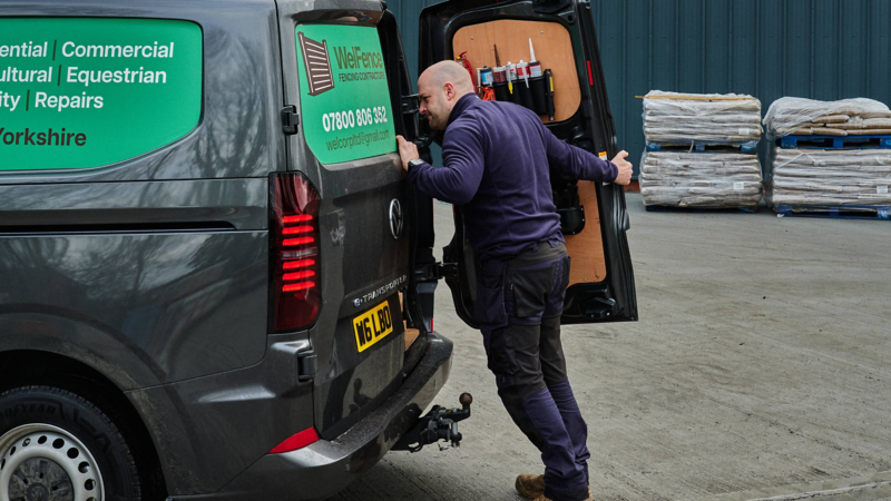 A man in work attire opens the back door of a gray van with a sign reading "Well-Fence." The scene is industrial, with pallets in the background.